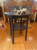 Front and side angled view of round wood occasional table standing on wooden floor with decorative items on top and behind on fireplace.