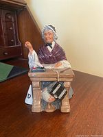 Front angle of seated elderly woman figurine depicting a schoolmarm behind a wooden desk with book and globe underneath.