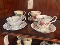 Four tea cups with matching saucers on wooden shelf, including floral designs and commemorative Canadian Centennial 1967 cup.