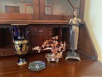 Photo showing entire lot: Greek vase, jade bonsai tree with cloisonné base, cloisonné dish, and metal statue on wooden sideboard.