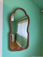 Full view of curvy-shaped wood framed wall mirror hanging on mint green wall, showing the frame's design and mirror glass reflecting a window with curtain and some furniture.