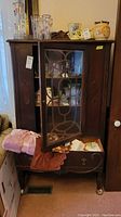 Front view of the vintage wood china cabinet showing upper glass door partially open, displaying glassware inside, bottom drawer partially open with fabric inside, wood carvings on sides and doors.