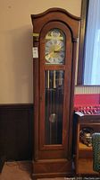 Full frontal image of the tall wooden grandfather clock showing the carved top, glass door, pendulum, and weights.