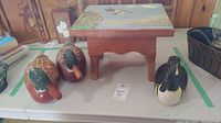 Two wood duck figurines and one wood duck head figurine displayed next to a painted wooden stool with coastal bird scene on seat.