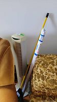 Photo showing tower fan, broom with yellow handle, and rolled window shade leaning against upholstered furniture.