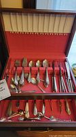 Wood box with red lining holding assorted plated forks, spoons, knives, and serving utensils in top section and bottom drawer.