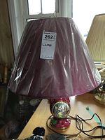 Full view of the lamp showing the purple-red plastic shade covered in protective plastic and the ceramic base with a painted decorative scene.