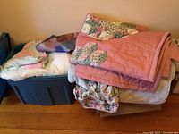 Stack of assorted linens including folded pink quilted blanket and floral patterned blankets placed on top of a closed dark gray plastic storage bin.