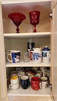 Two lower shelves containing assorted ceramic mugs with holiday themes, floral designs, and plain colors in the same cabinet.