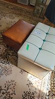 Photo showing a brown vinyl stool with storage next to a vintage cream vinyl tufted ottoman with green buttons on a patterned rug.