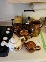 View of all tea pots, coffee pots, and stoneware pitcher on kitchen counter showing various colors and styles