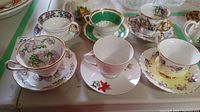 Six assorted porcelain teacups with saucers and a small plain white pitcher, displayed together on a table.
