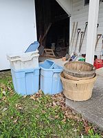 Plastic storage totes stacked with blue lids, showing dirt and outdoor use, next to planter baskets and gardening tools.