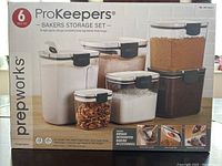 Front view of the boxed ProKeepers bakers storage set showing 6 labelled clear containers with black locking lids filled with various baking ingredients.