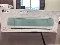Front of sealed retail box showing mint green Cricut Explore Air 2 machine illustration, brand logo, model name, and “2× Faster Cutting & Writing” text