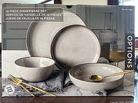 Boxed 16-piece Options dinnerware set featuring plates and bowls in speckled grey finish, shown with gold flatware and a textured grey cloth.