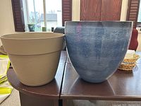 Photo showing two large flower pots placed on a wooden table indoors. Beige pot with texture on left, blue glossy pot on right.