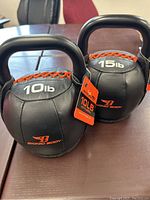 Photo showing two kettlebells with weight labels 10 lb and 15 lb, black body with orange stitching, and manufacturer tags attached.