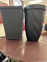 Photo of two large black plastic planters side by side on a brown table under natural light. One planter is cylindrical with herringbone texture and wide top rim, the other is rectangular with woven texture and tapering sides.