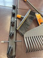 Top view of plastic storage hanger, broom attachment with black bristles, and leaf rake attachment with wide plastic tines. Items placed on brown surface.