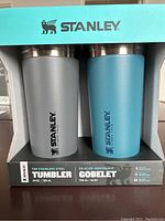 Front view of the 2 Stanley tumblers in grey and blue in original packaging with brand and product details visible.