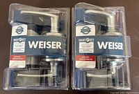 Front view of two new Weiser Smart Keyed Entry Door Handles in retail packaging showing chrome lever handles and keyway.