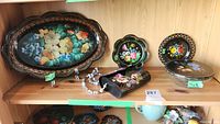 Shelf display showing two plates, a decorative bowl with a lid, and a floral purse with beaded strap all hand painted on black backgrounds.