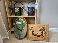 View of two hand painted metal watering cans on shelf, a wicker tray with painted tulips leaning on shelf, and a wooden laptop tray with painted floral design propped against wall.