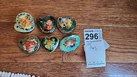 All six hand-painted floral brooches arranged on wooden surface, showing varied shapes and colors, with auction tag visible.