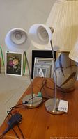 Two flexible shaft lamps with round metal bases and white shades on wooden table next to table lamp and framed pictures in background.