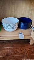 Two bowls on wooden shelf: white enamel mushroom-patterned bowl and solid dark blue ceramic bowl side by side