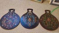 Three wooden Norwegian lefse boards side by side on carpet, each with distinct floral hand-painted folk art designs in varying blue and green shades with heart-shaped handle cutouts.