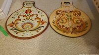 Both wooden Norwegian pancake boards side by side showing colorful hand painted floral and scroll patterns with heart-shaped handles.