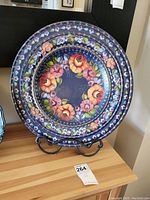 Full view of a round wooden plate hand-painted with Russian folk art floral designs and displayed on a metal stand.
