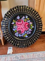 Full view of the hand-painted wooden plate featuring pink roses and gold detailing on a black background with scalloped edges, displayed on a black metal stand.