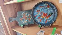 Both painted wooden items together on display: round plate and rectangular serving board with handle, showing floral designs and overall style.