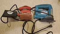 Makita 4323 jigsaw and King Canada 8351 sander side by side on carpet surface showing cords and dust collection bag on sander.