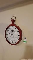 Clock hanging on beige wall showing full front view with round metal red frame and loop handle on top.
