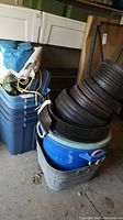 Five stacked blue Rubbermaid Roughneck bins with tarps and ropes on top, black plastic round planters stacked inside a blue bucket, galvanized bucket and blue muck bucket stacked together on concrete floor in garage