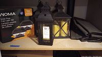 Photo of Noma Solar Post Cap Light Set box with five decorative lanterns and two outdoor curtains on shelf.