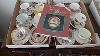 Long shot of two boxes containing twelve cups and saucers with the collectible book on top, showing variation in patterns and colors.