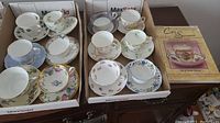 Twelve cups and saucers displayed in two boxes with floral and gold designs, included collectible book