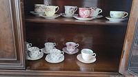 Full view of twelve assorted floral patterned cups and saucers on two shelves inside wooden cabinet