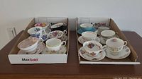 Twelve assorted bone china cups and saucers arranged in two boxes showing various floral designs and gold trims.