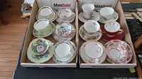 Full set of twelve assorted cups and saucers displayed in two boxes, showing various floral and decorative patterns with gold trims.