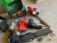 Scotts fertilizer spreader shown with folded handle, electric blower vacuum with collection bag, and Black & Decker hedge trimmer laying on floor.