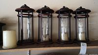 Four metal framed lanterns with cylindrical glass sleeves arranged side by side, showing overall condition and style.