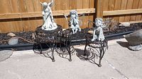 Three outdoor garden figures including one large angel and two smaller fairies displayed on three decorative metal tables against a wooden fence.