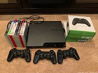 PS3 console with three black controllers, eight PS3 games stacked beside it, two Xbox controllers in boxes included