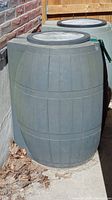 Two plastic rain barrels shown outside next to a brick wall, both have a round lid.